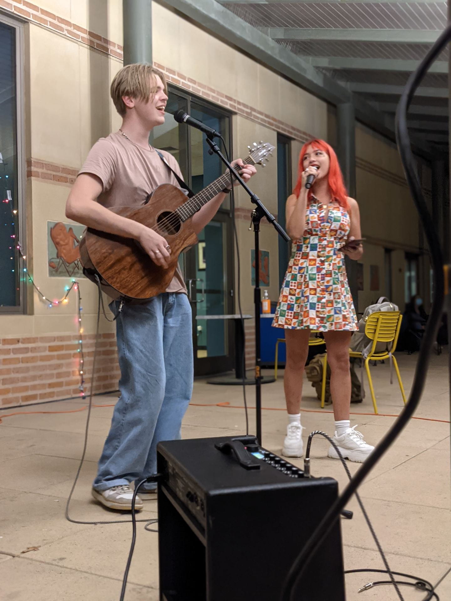 A picture of two college students, a man and a woman, performing a song at an outdoor college event. The man is playing an accoustic guitar, and the woman is singing into a microphone